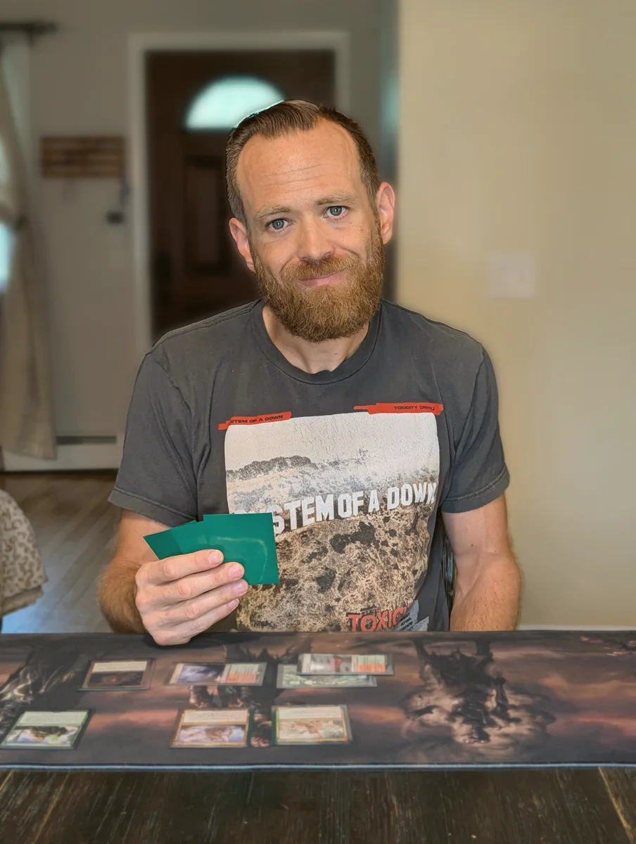 Chris Coyle at his kitchen table mid-game, smiling, holding a fanned hand of sleeved Magic cards over a playmat